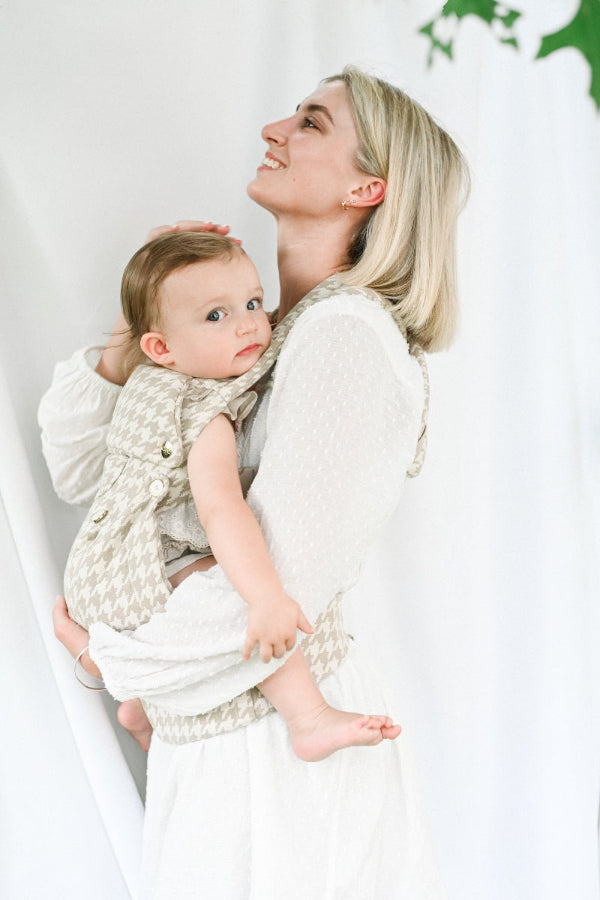 A radiant blonde mother in a textured white "Swiss dot" blouse smiles upward in profile against a soft white backdrop. She holds a blue-eyed toddler in a beige houndstooth carrier with gold button accents - the Grace Kaya. The baby looks calmly at the camera while bare feet dangle. The image is bright, airy, and exudes elegant joy.