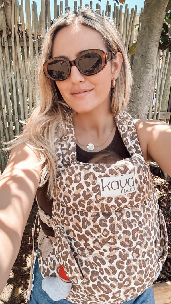 A sun-drenched selfie of a smiling blonde woman in oval tortoiseshell sunglasses. She wears a leopard-print Kaya Baba, the Savvanah, on her chest holding an infant - only a tiny grey-socked foot is visible. Spotted leaf shadows fall across her and the rustic wooden fence background. The vibe is warm and summery and very luxurious.