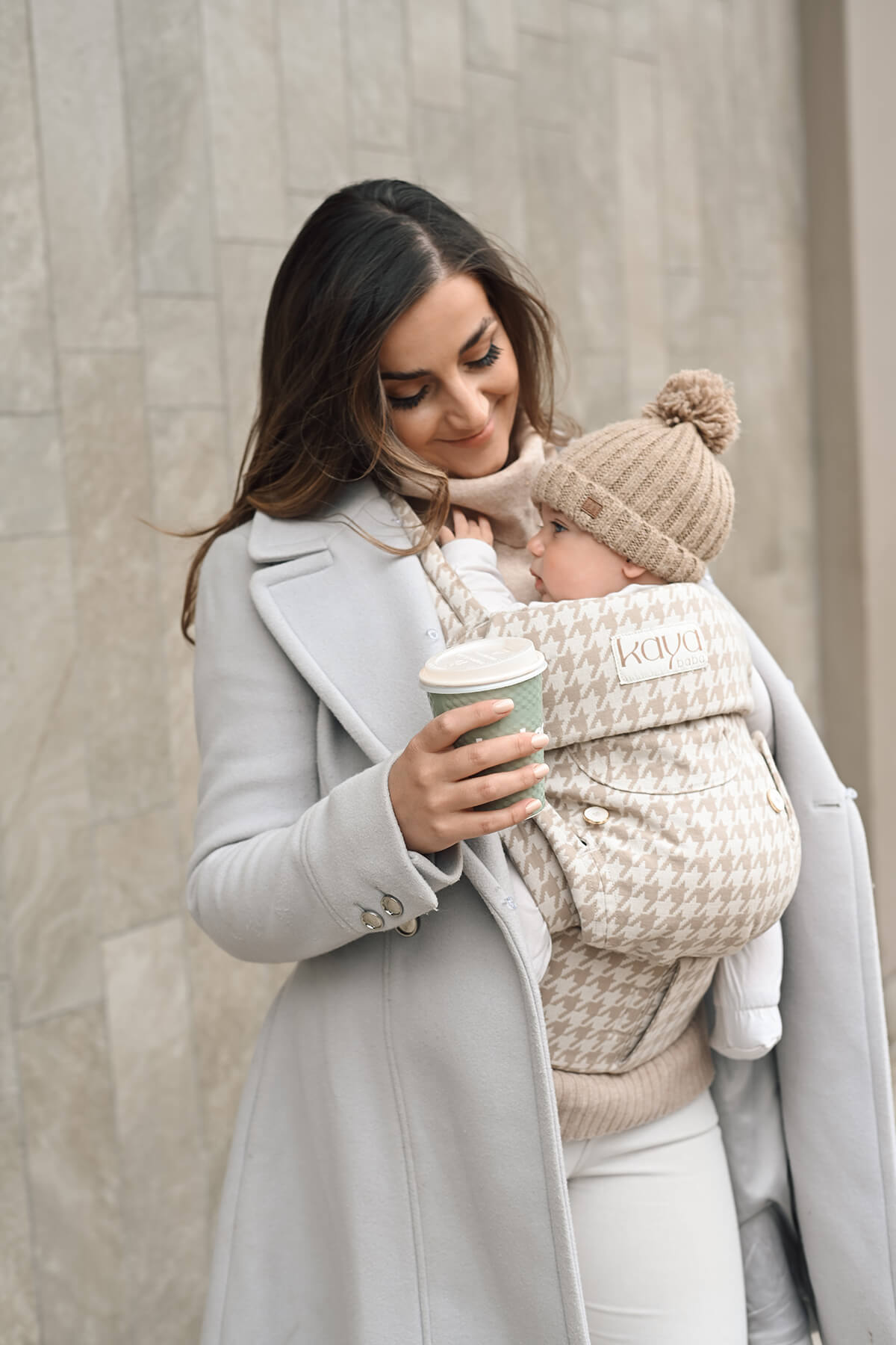 A dark haired woman with a gray coat is wearing a beige houndstooth Kaya, the Grace Kaya, with a baby boy inside. The boy is wearing a brown wool beanie. The woman is is holding a coffee in her right hand while lovingly looking at her baby boy. The background is a marble brown tiled wall.