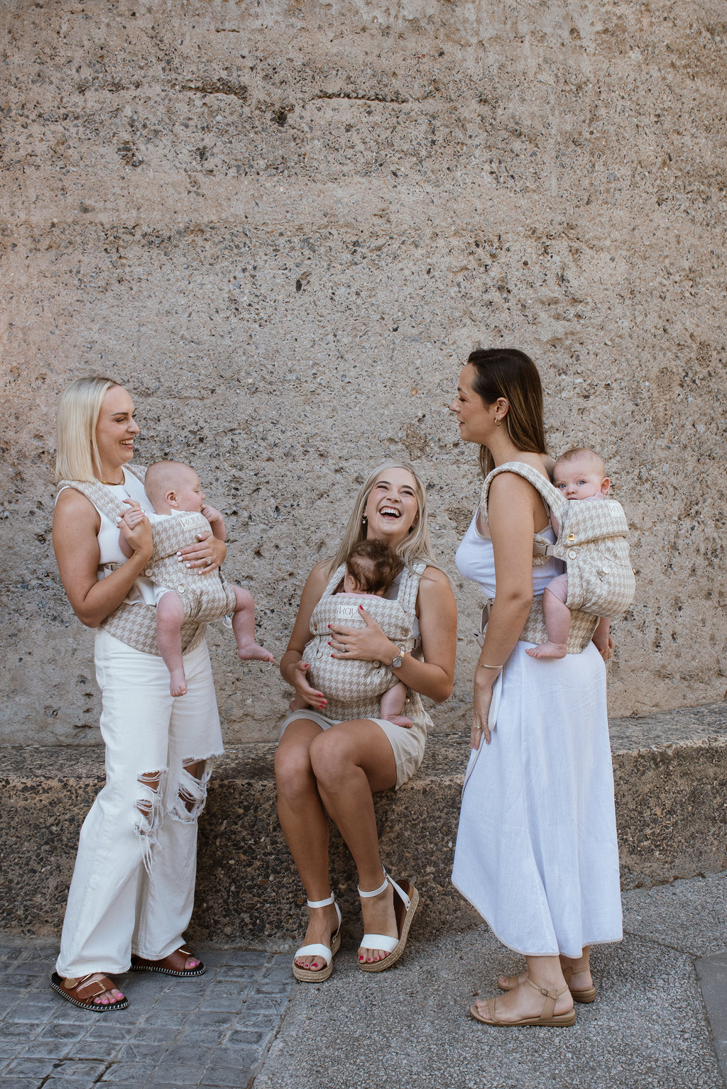 Three caucasian women standing in front of the Silo Hotel in Cape Town all wearing Grace Kaya's, the beige houndstooth Kaya, and carrying their babies in three different positions: On the back, front forward facing and front inward facing. All of the Moms are wearing white clothing and are laughing and smiling.