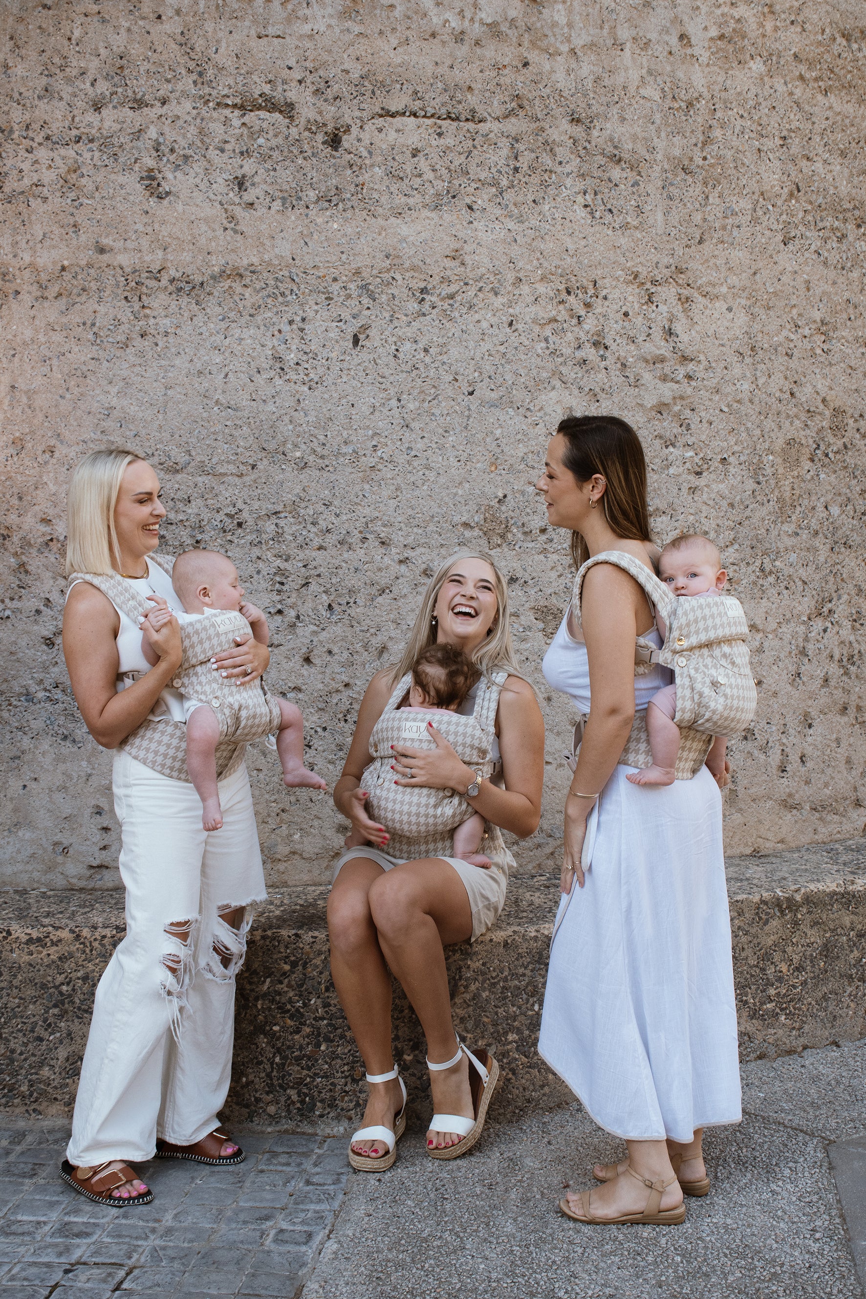 Three caucasian women standing in front of the Silo Hotel in Cape Town all wearing Grace Kaya's, the beige houndstooth Kaya, and carrying their babies in three different positions: On the back, front forward facing and front inward facing. All of the Moms are wearing white clothing and are laughing and smiling.