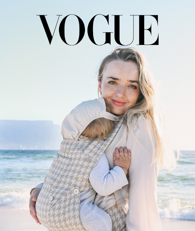 Woman holding a child in a baby carrier on a beach with 'Vogue' magazine cover text.