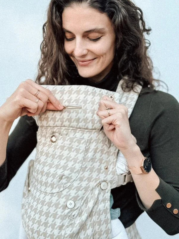 A close-up of a curly-haired woman in a dark turtleneck adjusting the zipper on a beige houndstooth baby carrier. She smiles softly downward, wearing gold rings and a watch. The focus is on the carrier's functional details and premium buttons against a plain light background.